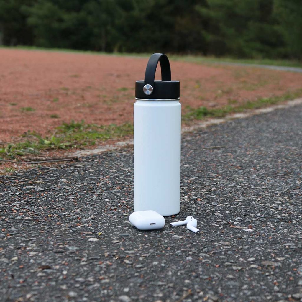 A white metal water bottle with a black lid is placed on the ground of a running track, with a pair of wireless headphones beside it.