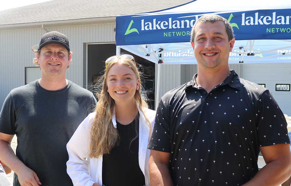 Three Lakeland Networks employees stand outside in front of a blue tent with the Lakeland Networks logo on it. From left to right, a young man in a black shirt and baseball cap, a young woman with long hair in a white shirt, and a young man in a black shirt. 