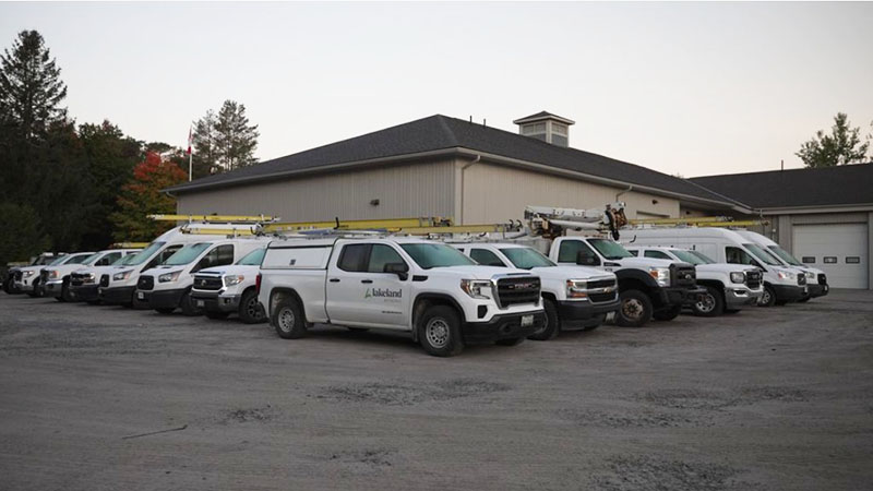 A fleet of white Lakeland Networks vehicles, including pickup trucks, vans and cars, in front of Lakeland's office in Bracebridge. 