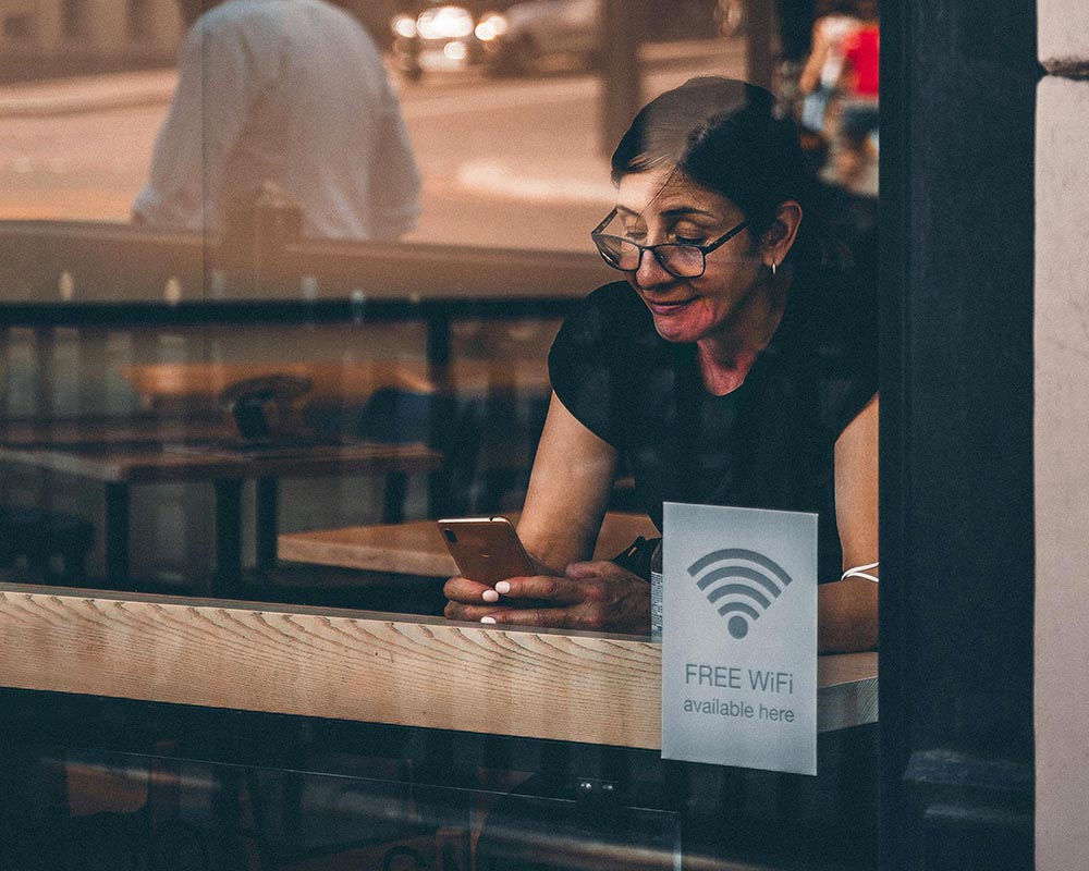 A dark haired woman with glasses sits at a counter behind a window, looking at her phone. A sign in the window reads "Free WiFi available here"