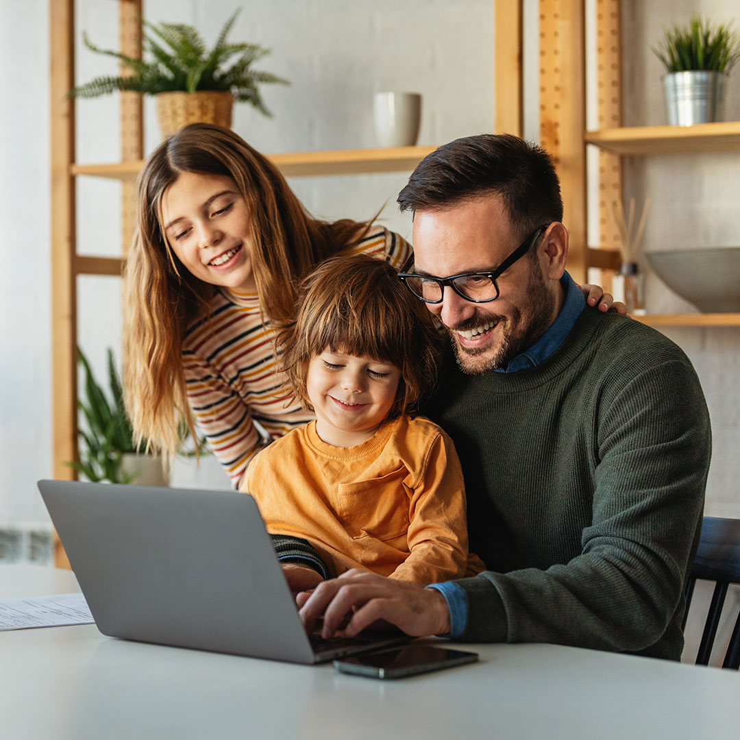 A father and two children sit in front of a laptop at a table